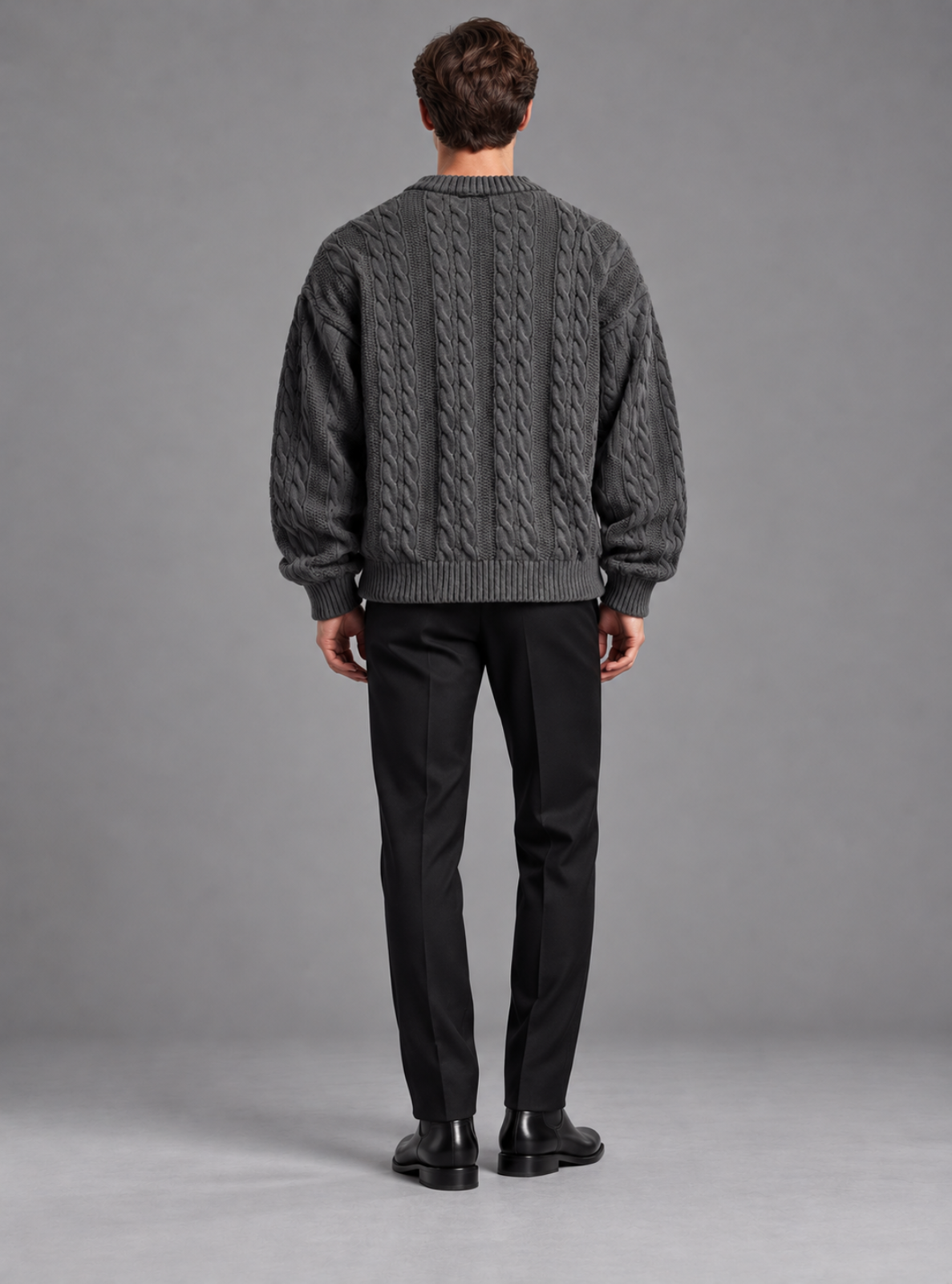 A man viewed from behind wearing a dark gray cable-knit sweater and black trousers, standing against a neutral gray studio background.
