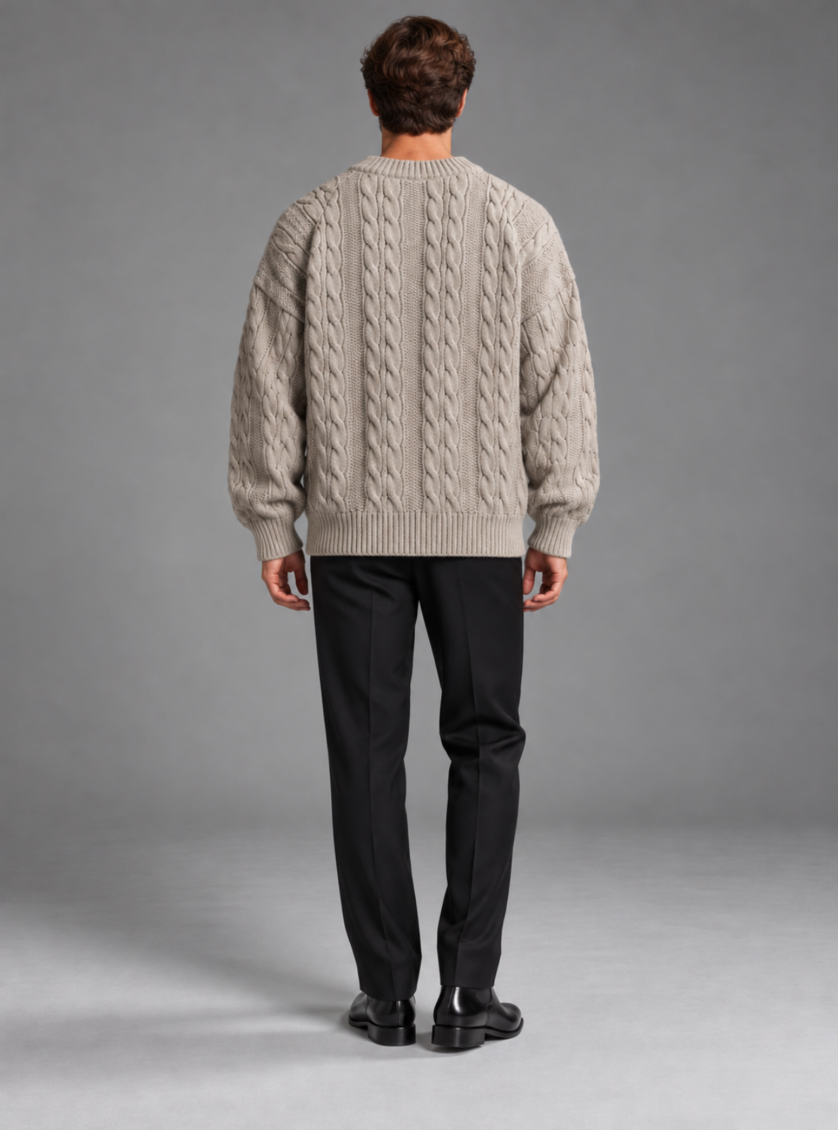 A man viewed from behind wearing a light beige cable-knit sweater, black trousers, and black shoes, standing against a neutral gray studio background.
