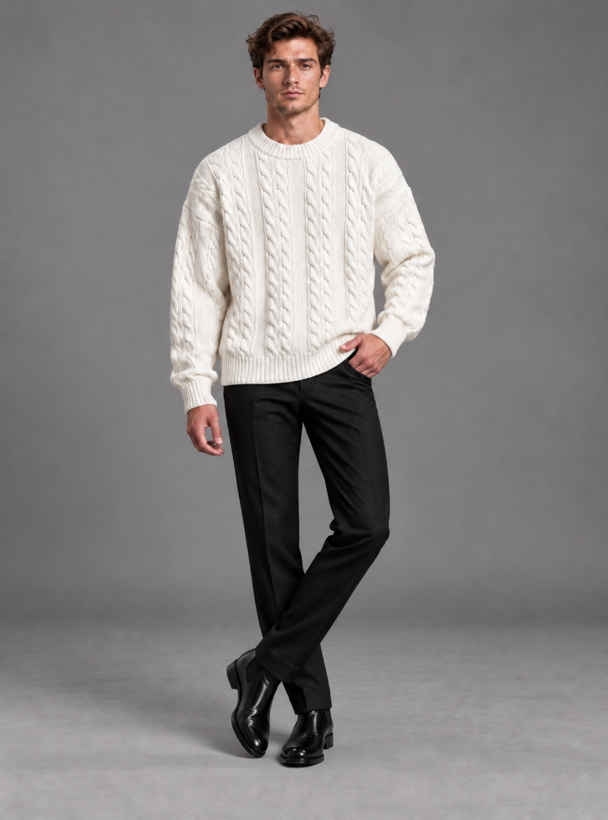 A man modeling a cream cable-knit crewneck sweater paired with black trousers and black leather dress shoes, standing against a solid gray studio background.
