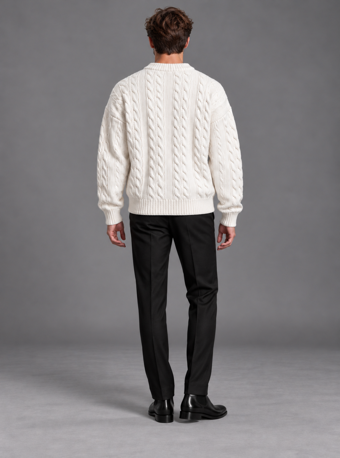 A man viewed from behind wearing a cream cable-knit sweater, black trousers, and black shoes against a plain gray studio background.