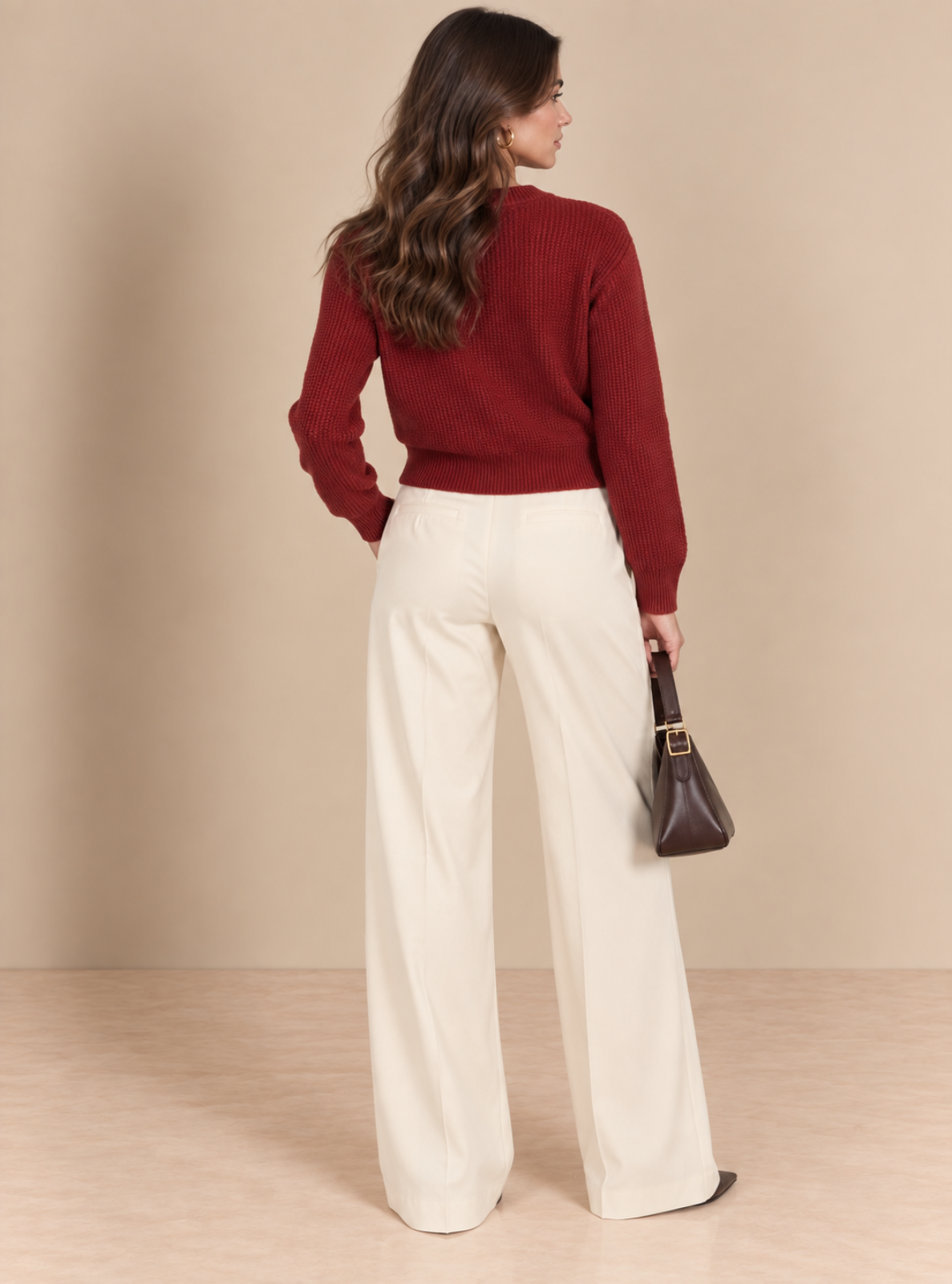 A woman is shown from behind wearing a dark red ribbed knit sweater and high-waisted off-white wide-leg trousers, holding a dark brown handbag against a neutral beige studio background.