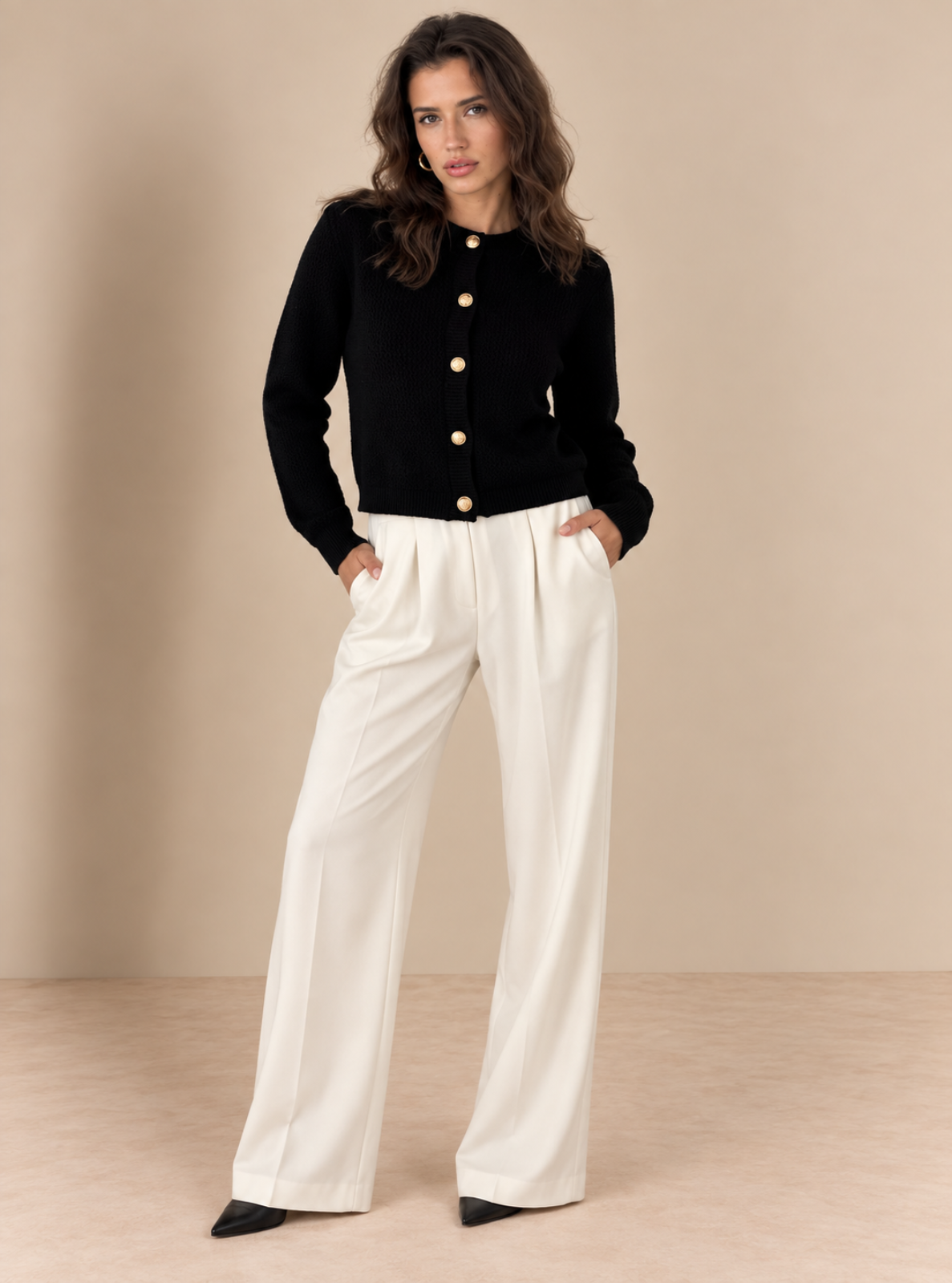 A woman modeling a black knit cardigan with gold buttons and white wide-leg trousers, standing against a neutral beige studio backdrop.