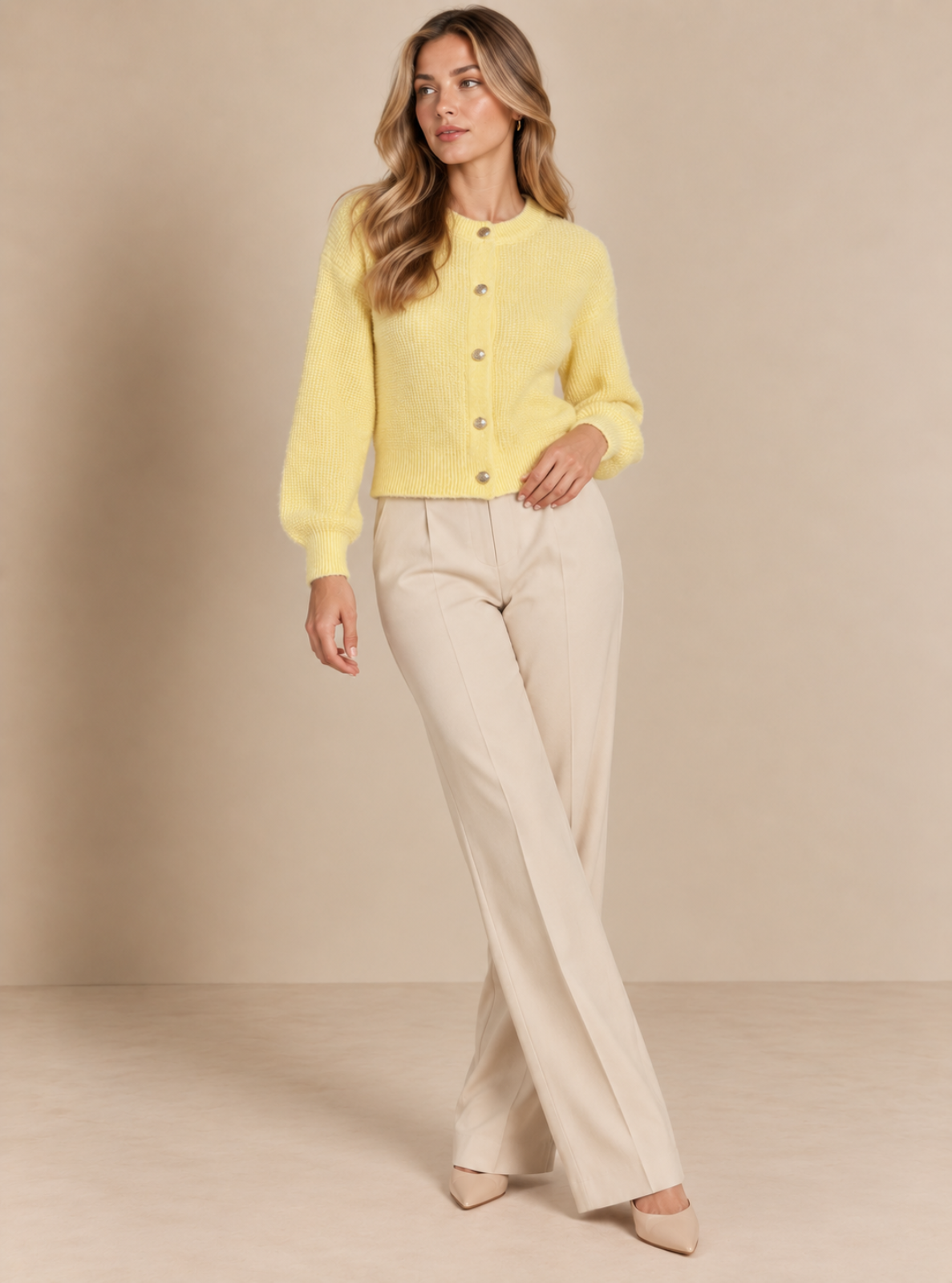 A woman modeling a pale yellow long-sleeve button-front cardigan sweater paired with cream-colored wide-leg trousers, standing against a solid beige studio backdrop.