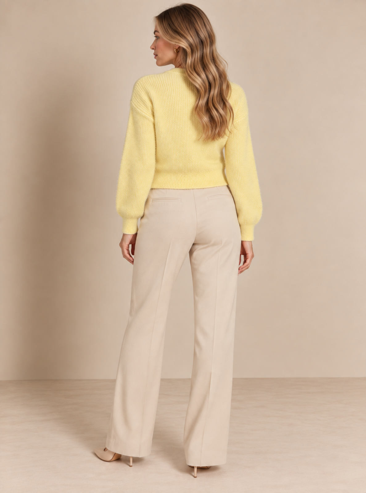 A woman shown from the back wearing a pale yellow knit sweater and light beige wide-leg trousers, standing against a plain beige studio backdrop.