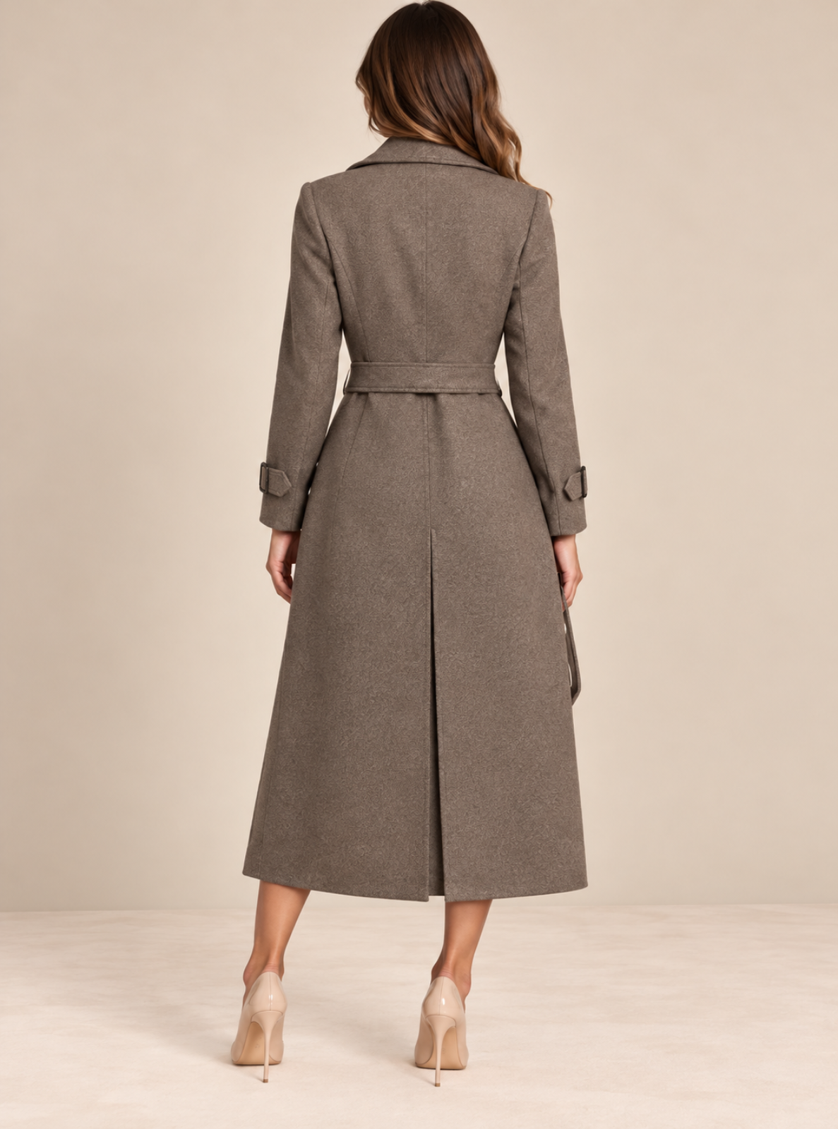 A woman viewed from the back wearing a long taupe-brown belted coat and beige heels, standing against a plain beige studio background.