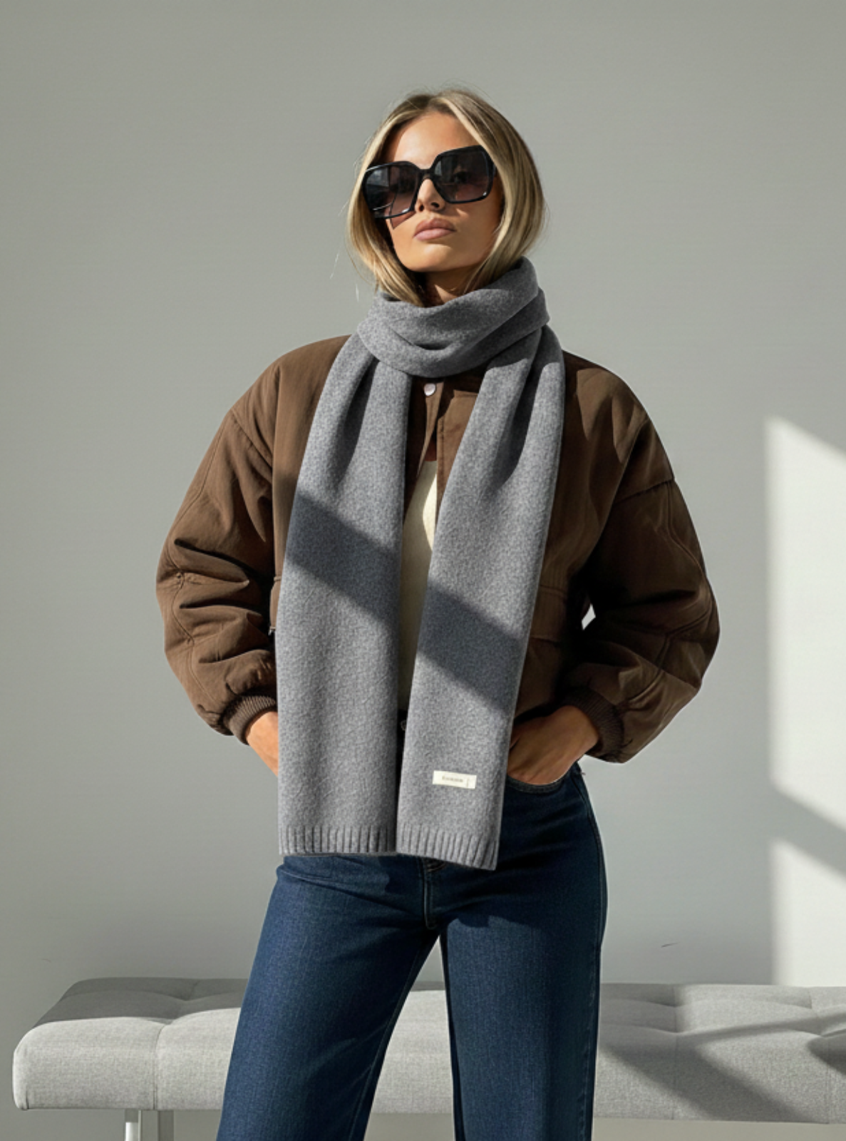 Woman wearing a brown coat, gray scarf, and sunglasses in a sunlit room.