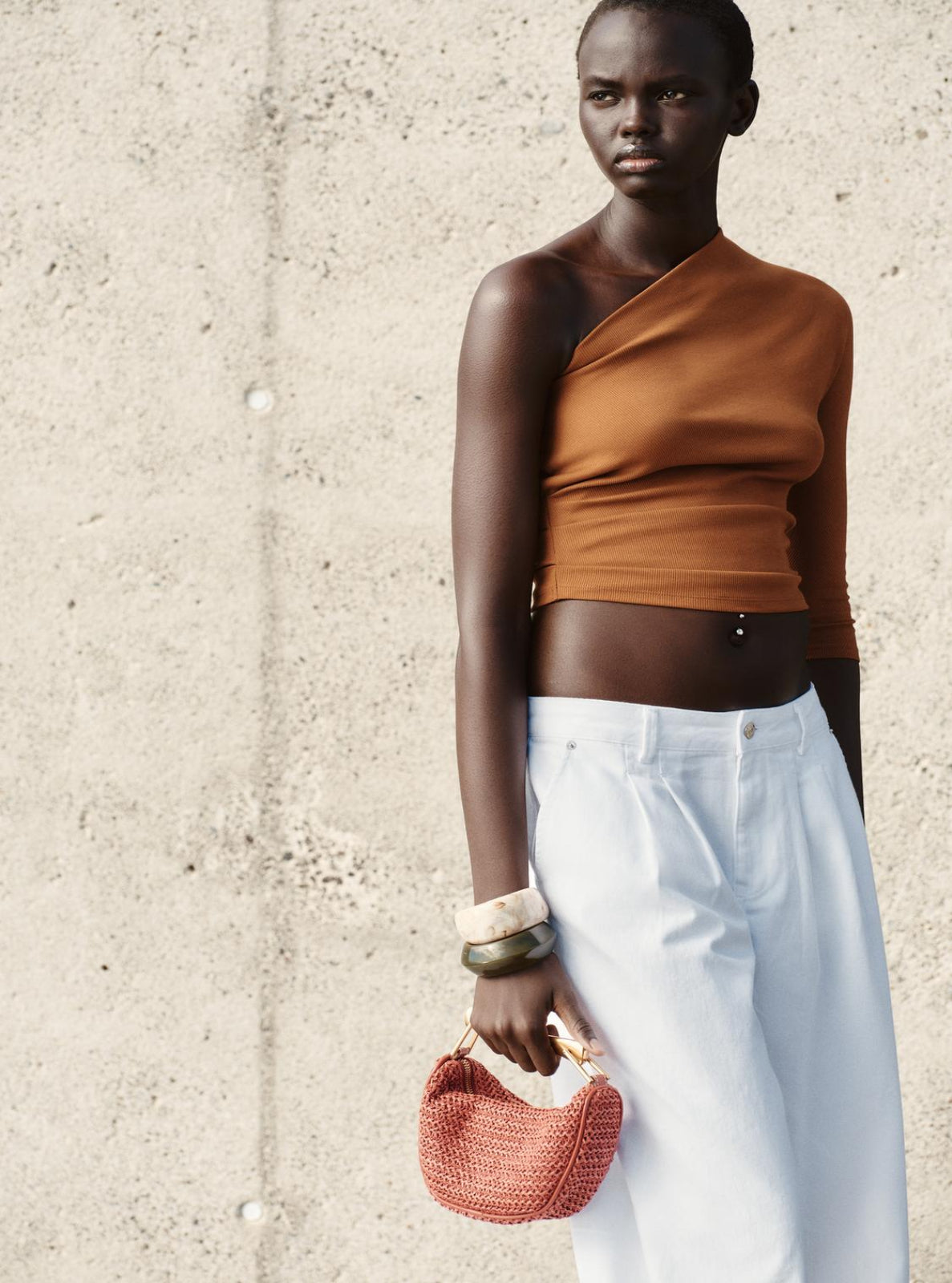 Woman wearing a brown top and white pants, holding a red handbag against a concrete wall.